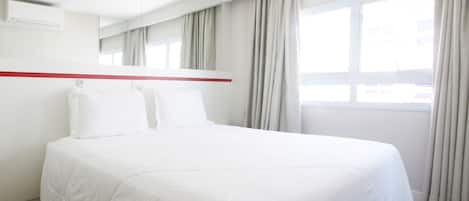 Una habitación de hotel moderna con una cama grande, un espejo, una pared de acento roja y un ventanal con cortinas.