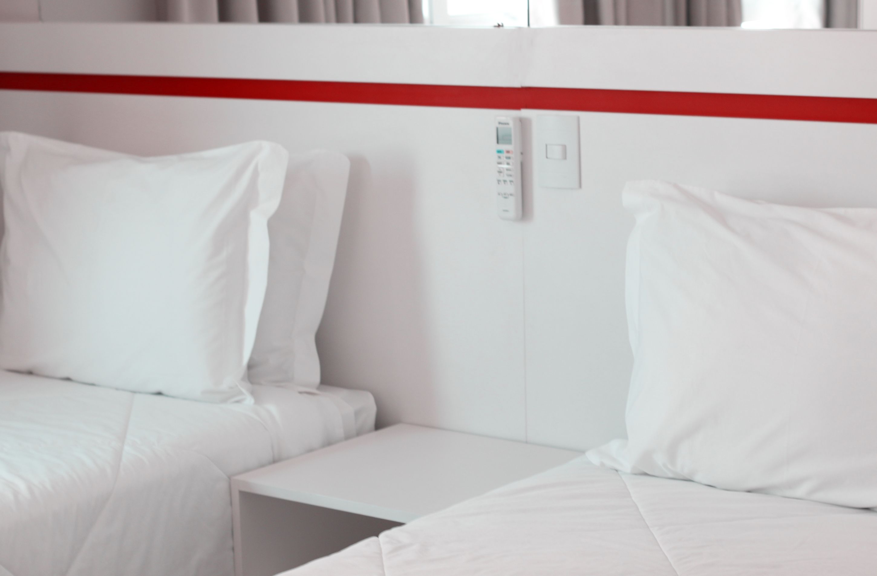 A hotel room with two beds, white bedding, a red accent wall, and a control panel on the wall.