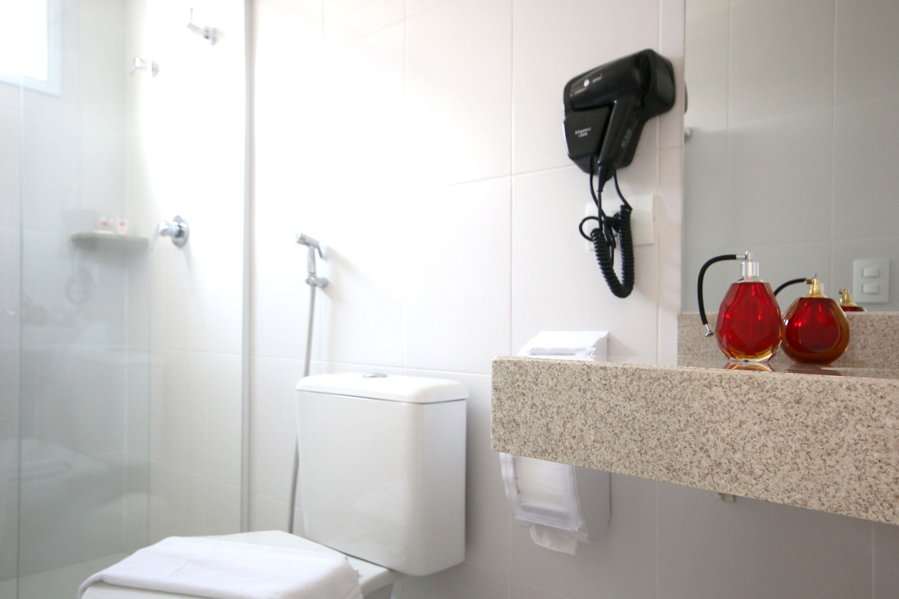 Casa de banho com cabine de duche em vidro, sanita branca, bancada com secador de cabelo e uma garrafa vermelha.