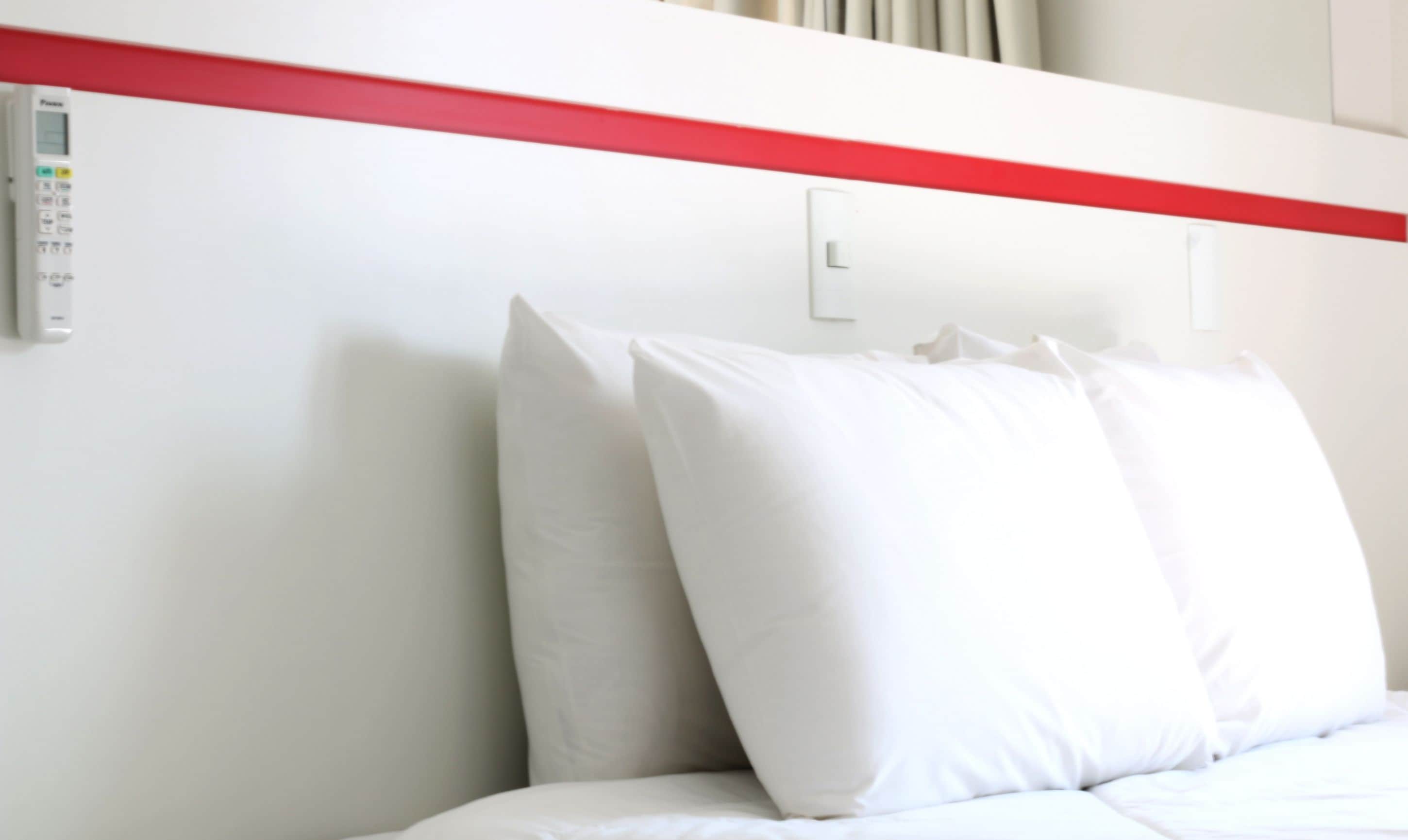 Ein Hotelzimmer mit Bett, weißen Kissen, einer roten Akzentwand und einem Thermostat an der Wand.