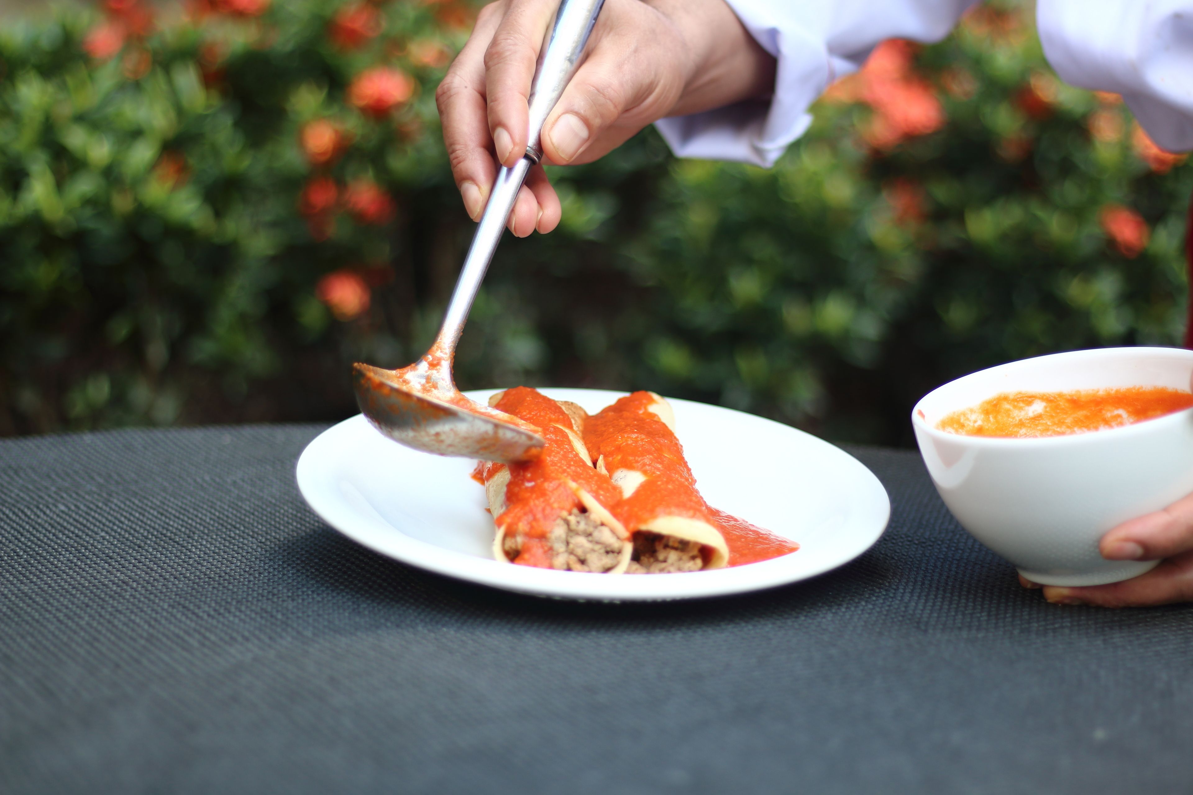 A person serving a dish with a spoon on a white plate, with a bowl of sauce nearby.