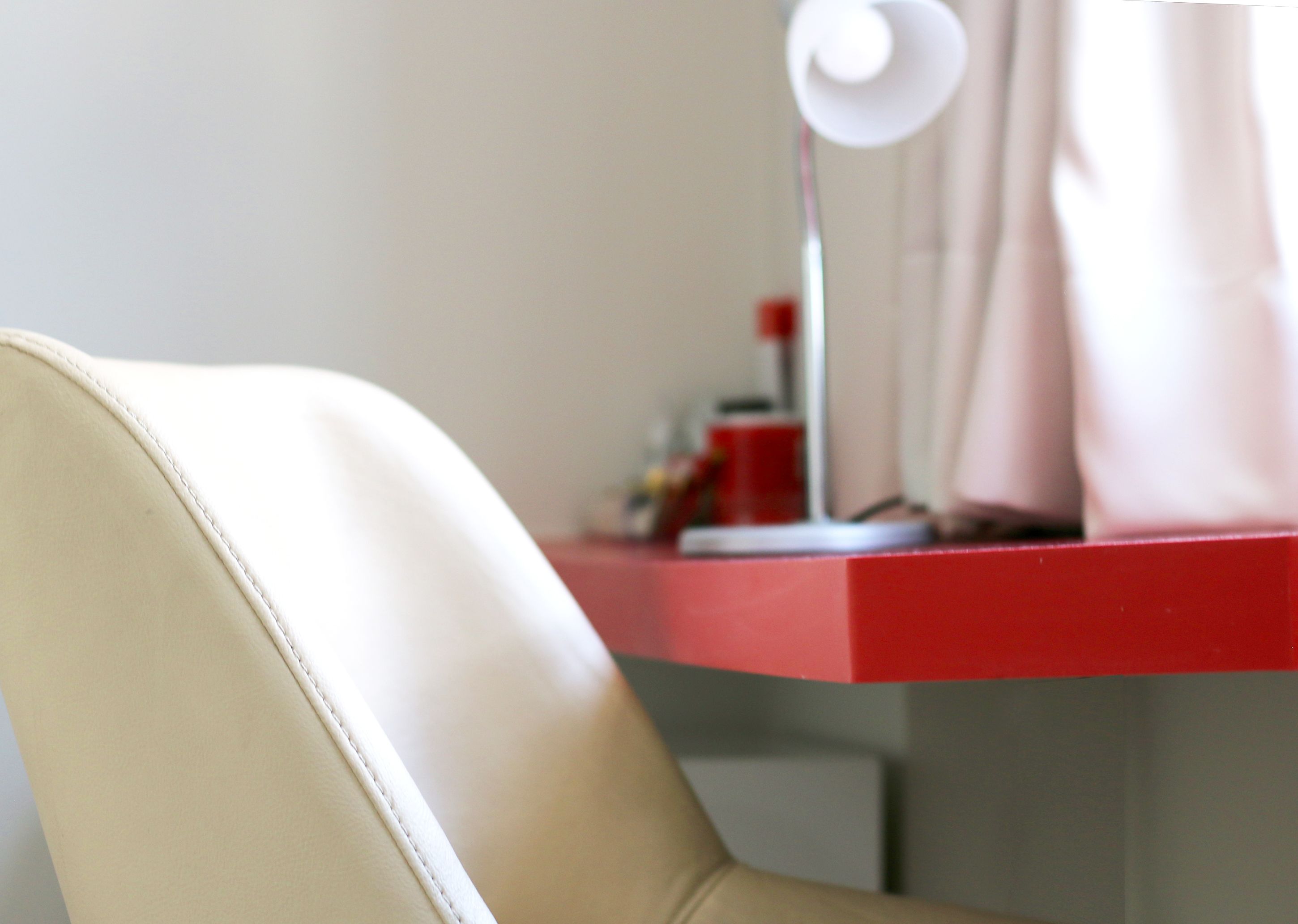 A close-up of a chair with a white leather backrest and a red desk with a white lamp.