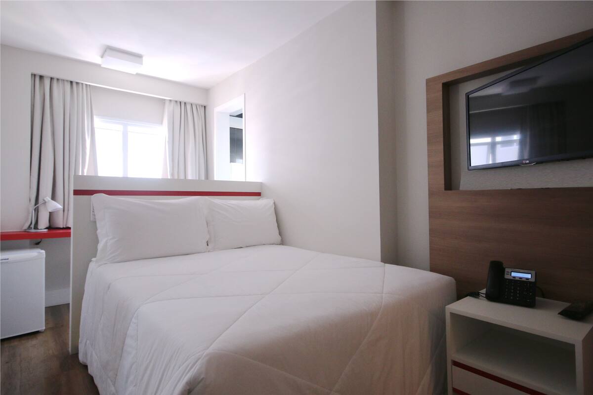 Une chambre d’hôtel avec un lit, un téléviseur fixé au mur, une table de chevet avec un téléphone et une fenêtre avec des rideaux.