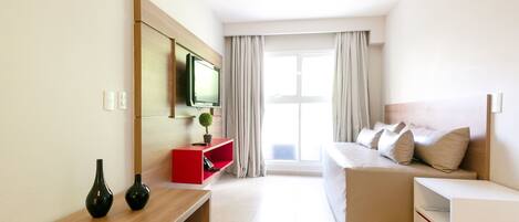 Una habitación de hotel moderna con una cama, un televisor montado en la pared, una mesita con dos jarrones, un mueble de almacenamiento rojo y un amplio ventanal con cortinas.