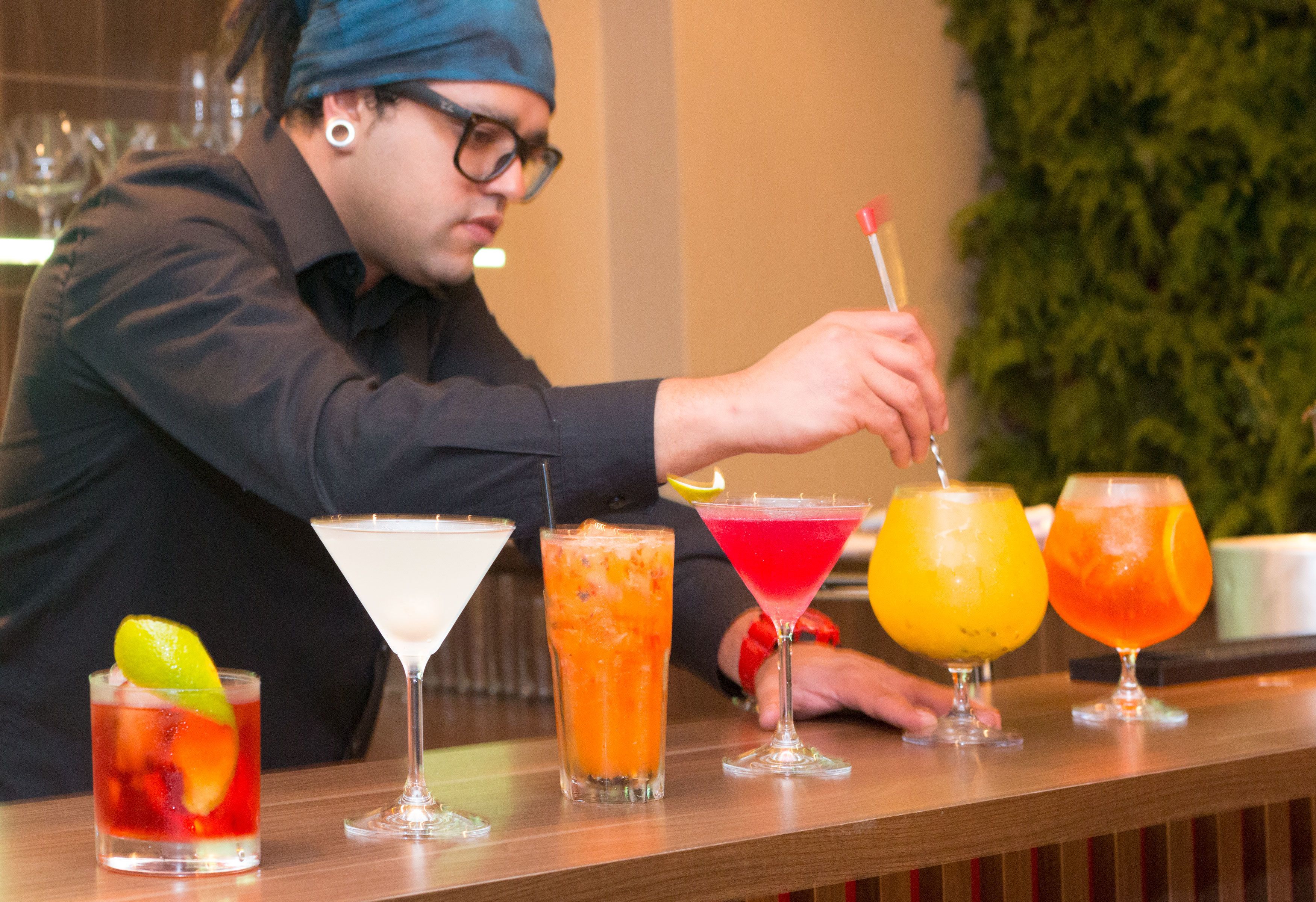 Un barman prépare des boissons dans un bar où l’on peut voir divers cocktails et une plante verte en arrière-plan.