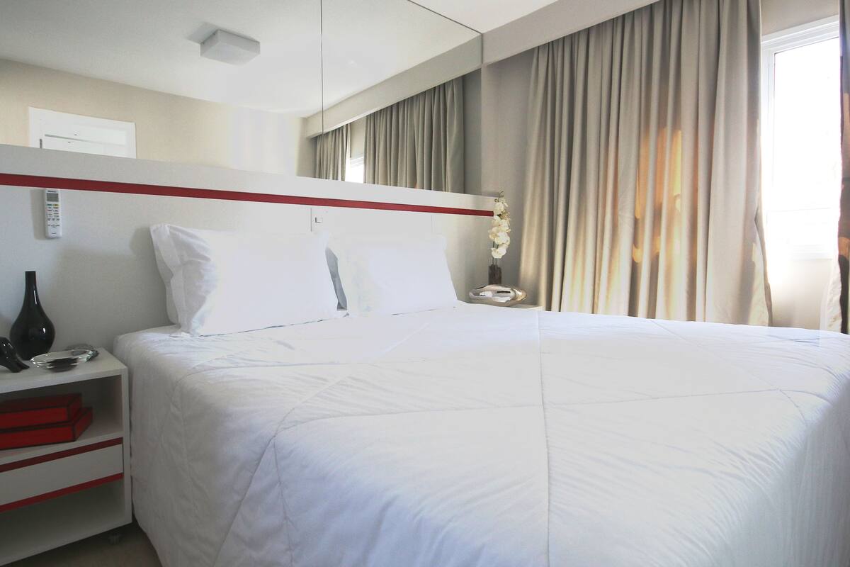 Une chambre d’hôtel avec un grand lit, une table de chevet avec un téléphone et un vase, un meuble de rangement rouge et une fenêtre avec des rideaux.