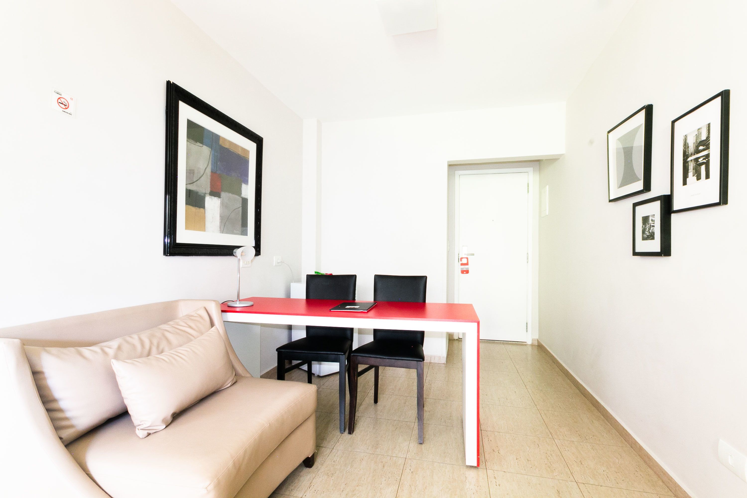 A modern living room with a beige sofa, a red and white table, and black chairs. There are framed pictures on the walls and a door in the background.