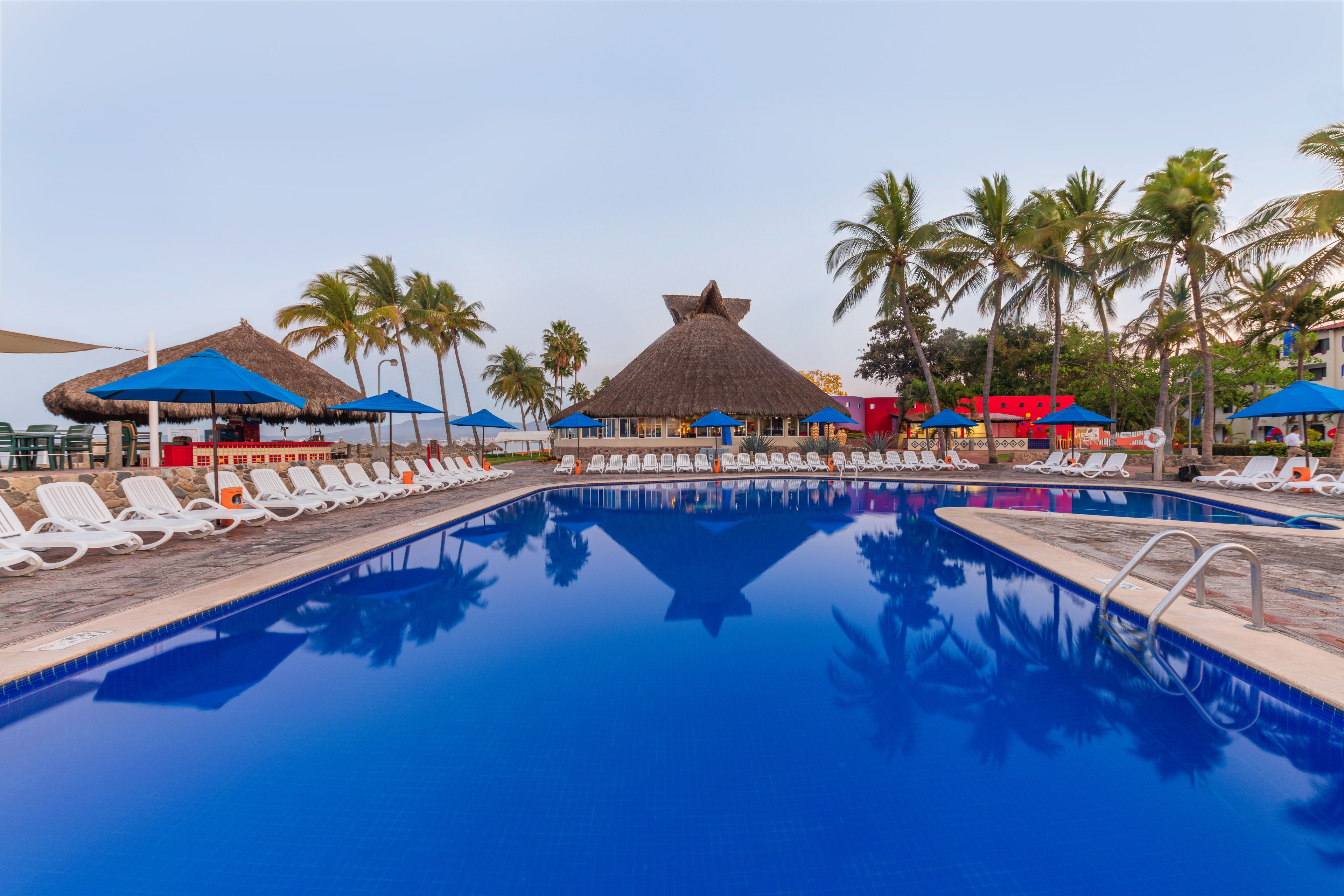 Ein Pool mit Liegestühlen und Sonnenschirmen, umgeben von Palmen und Gebäuden.
