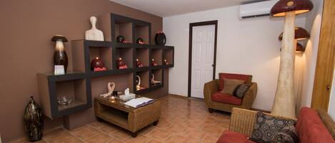 A living room with a brown sofa, a red sofa, a wicker coffee table, and a wall-mounted shelf with decorative items.