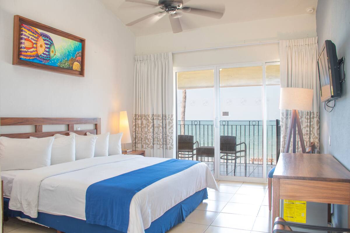 A hotel room with a bed, a ceiling fan, a balcony with chairs, and a painting on the wall.