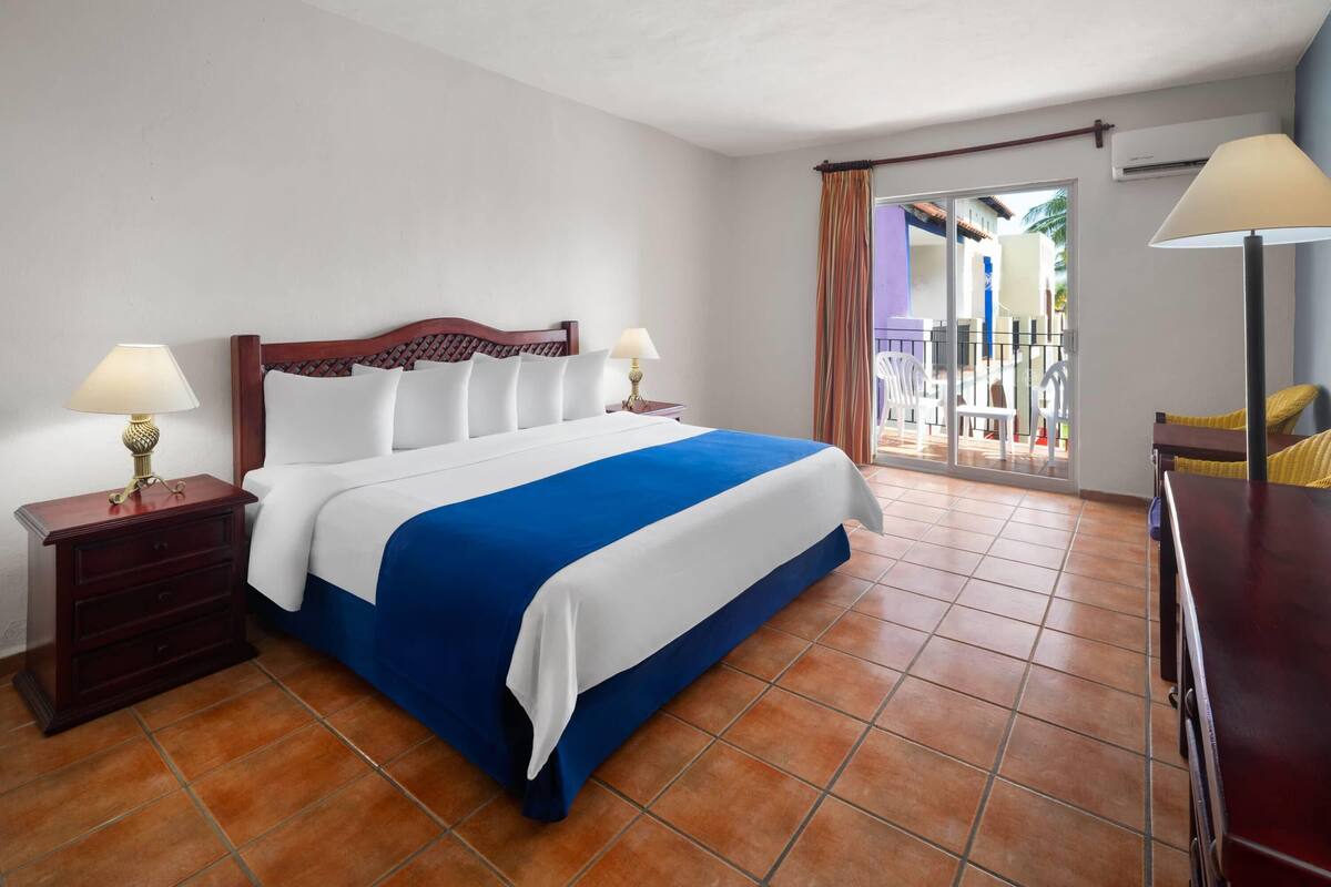 A hotel room with a bed, two bedside tables with lamps, a wooden headboard, a blue and white bedspread, a red dresser, and a view of the outside balcony.