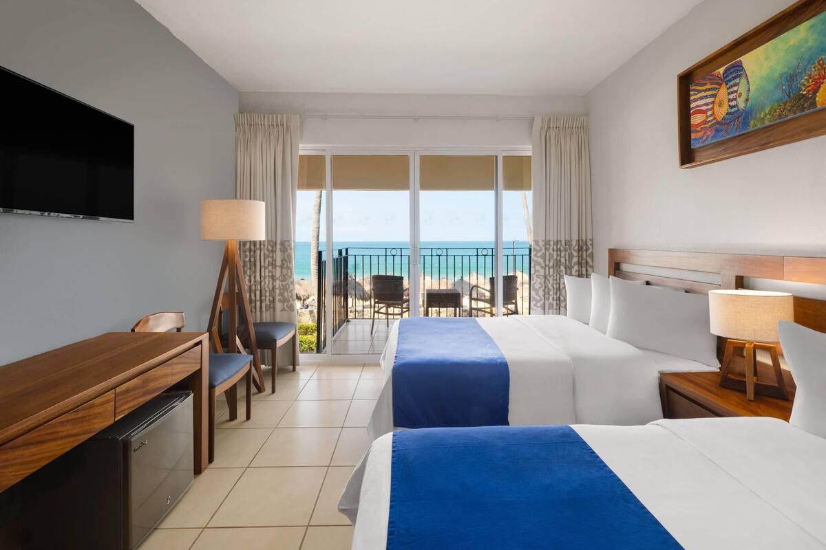 A hotel room with a bed, a desk, a television, and a balcony with a view of the ocean.