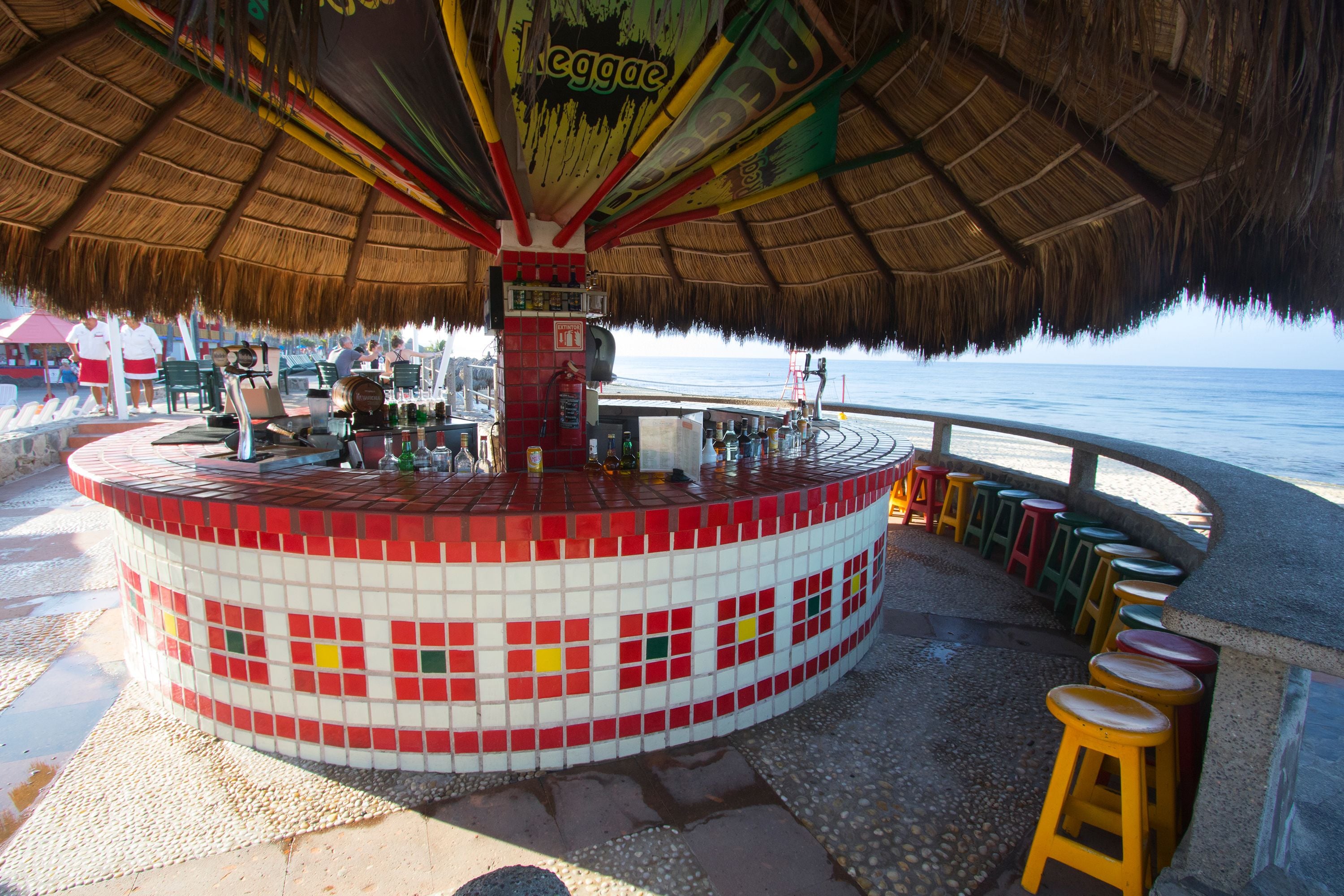 A beachside bar with a tiled exterior, a thatched roof, and stools along the edge.
