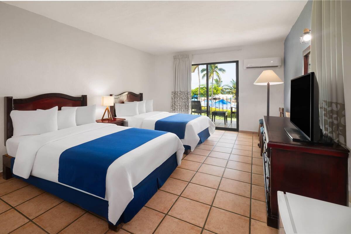 A hotel room with two beds, a TV, a desk, and a view of the outdoors.