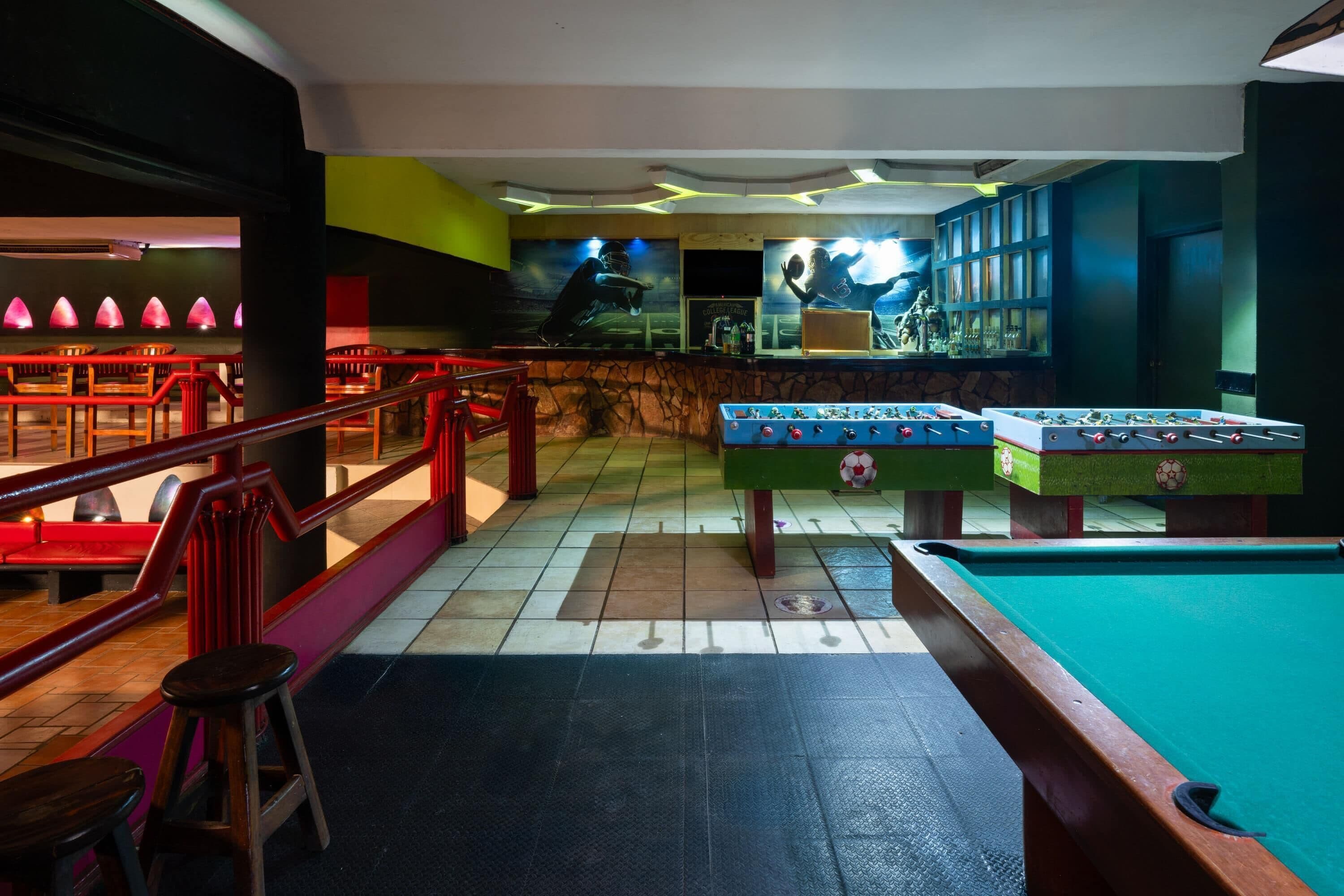 A billiards room with two foosball tables, a pool table, and a bar area with a mural.