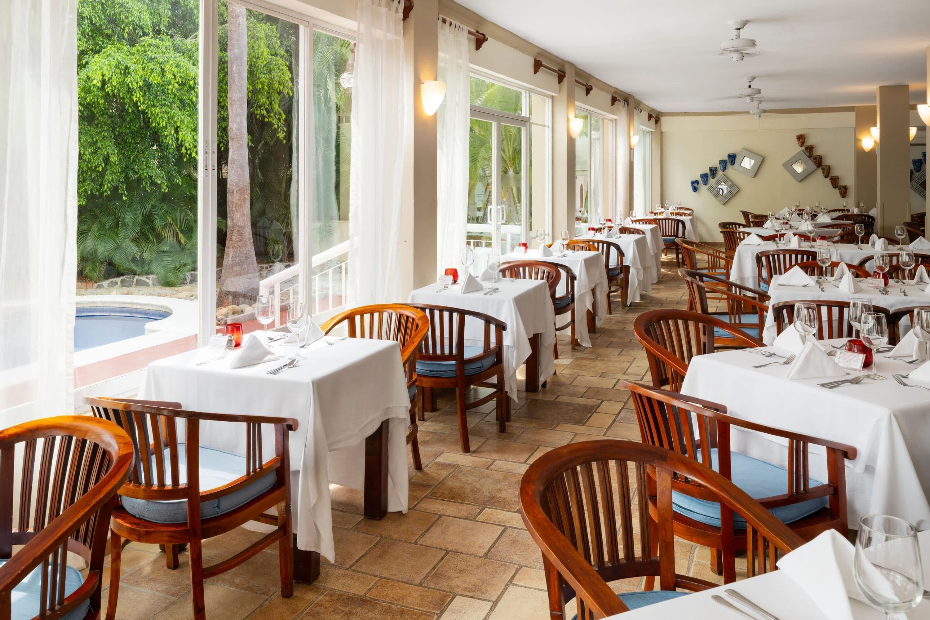 Un restaurant doté de nombreuses tables et chaises, de grandes fenêtres avec des rideaux et d’un sol carrelé.