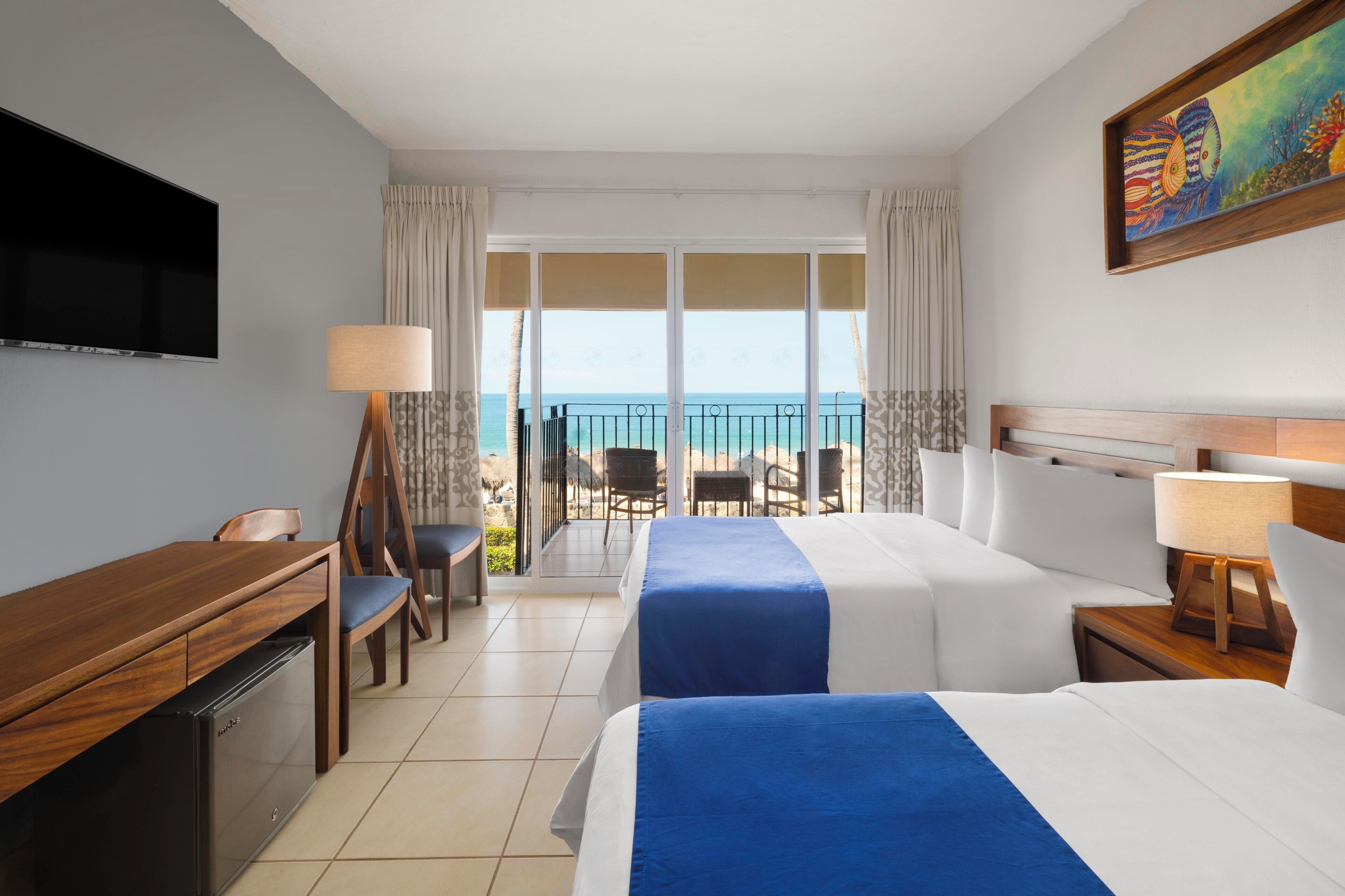 A hotel room with a TV, a desk, a chair, two beds, a balcony with a view of the sea, and a painting on the wall.