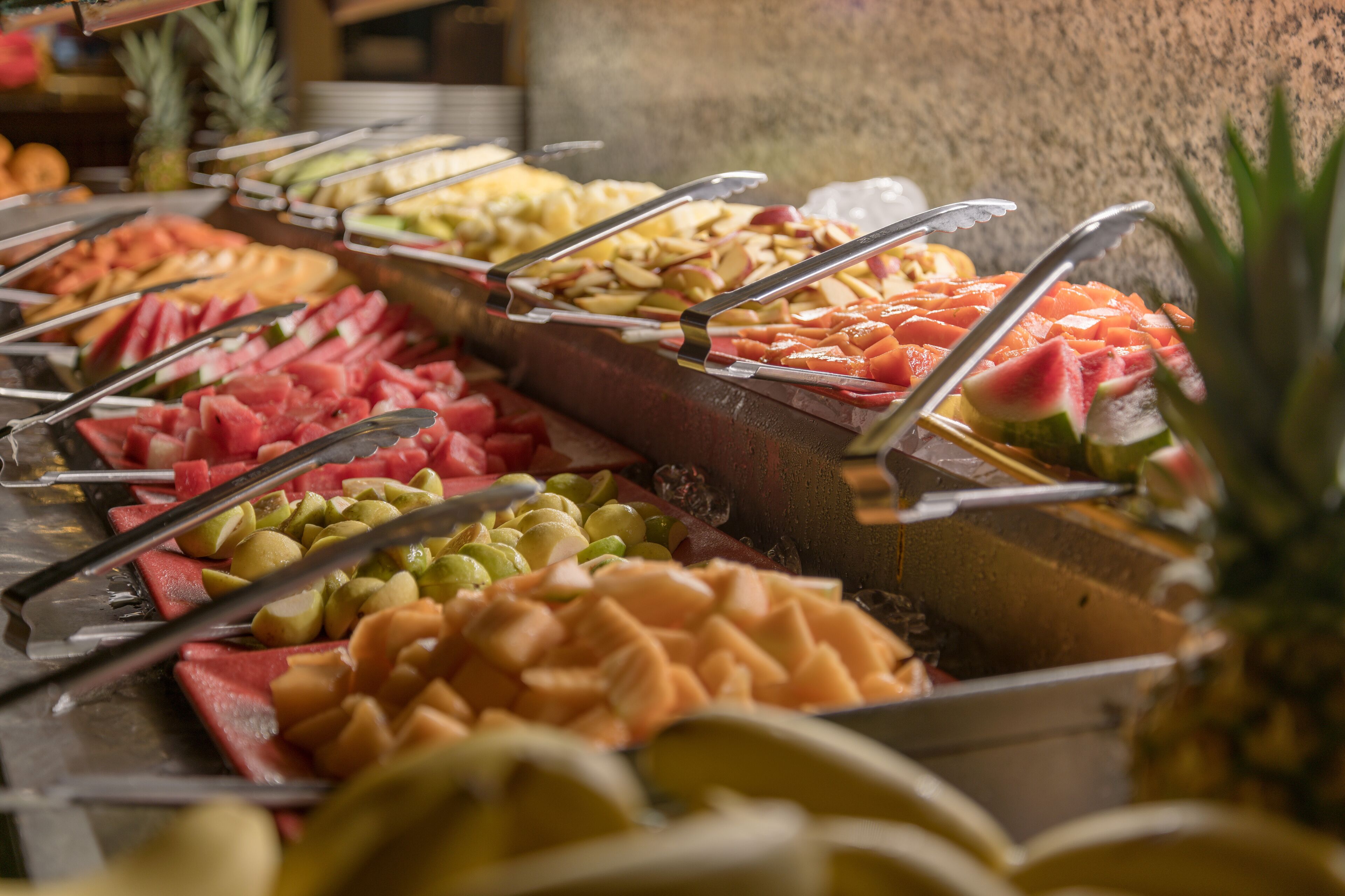 Um buffet com diversas frutas e legumes dispostos em recipientes separados, cada um com pinças para servir.