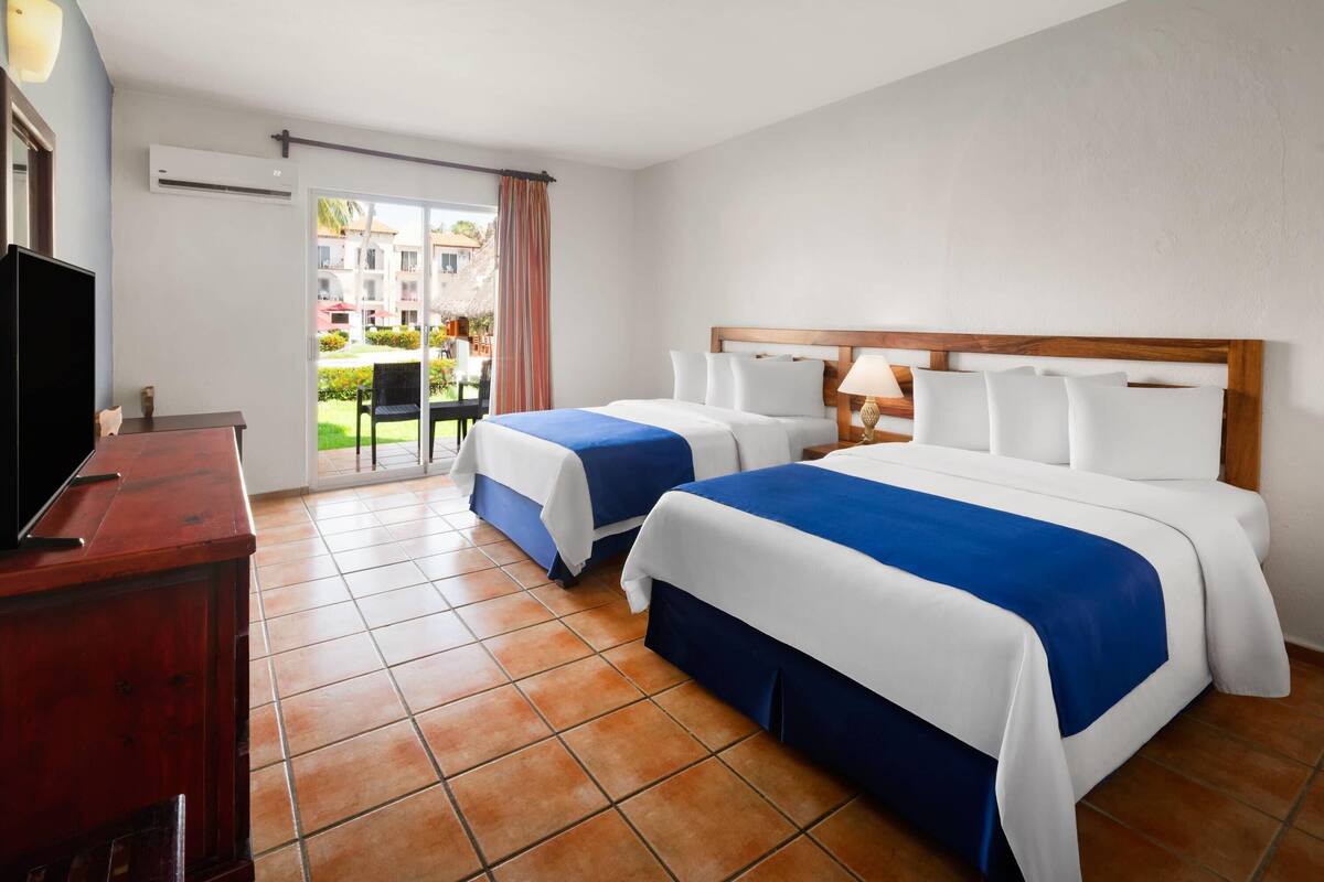 A hotel room with two beds, a TV, a desk, and a balcony with a view of buildings and greenery.