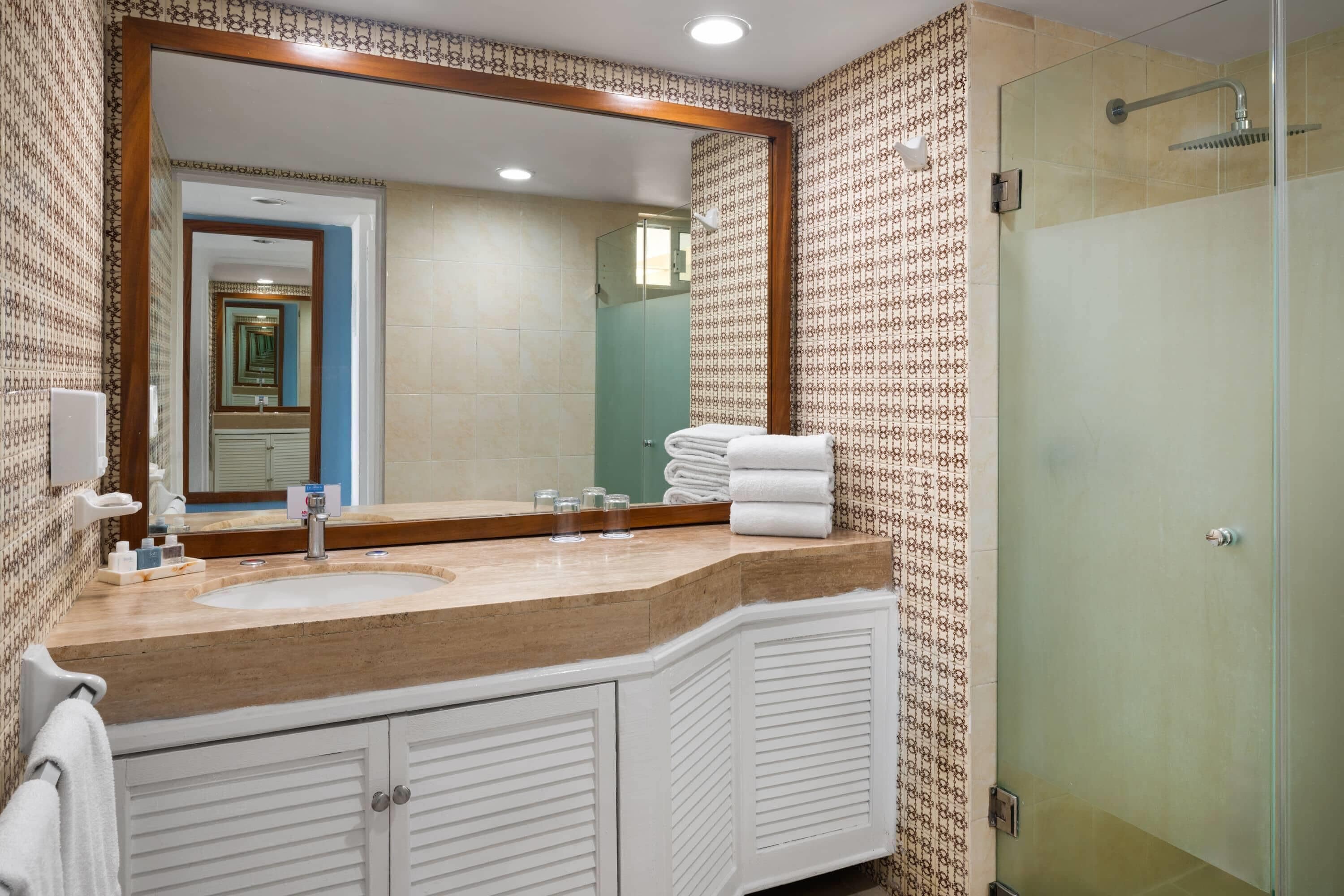 A bathroom with a large mirror, a sink, a shower, and patterned wallpaper.