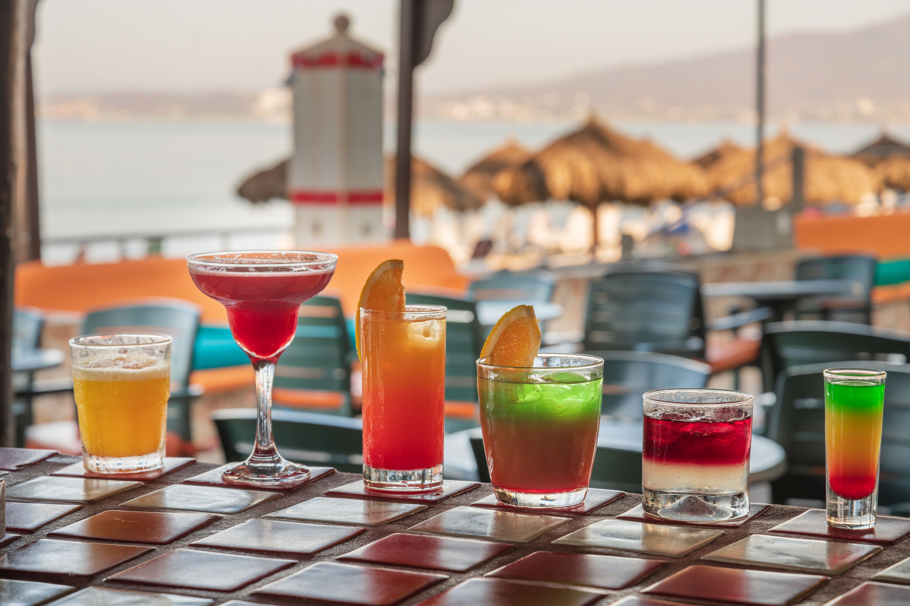 Una mesa con diversas bebidas de colores, un faro al fondo y sombrillas de playa.