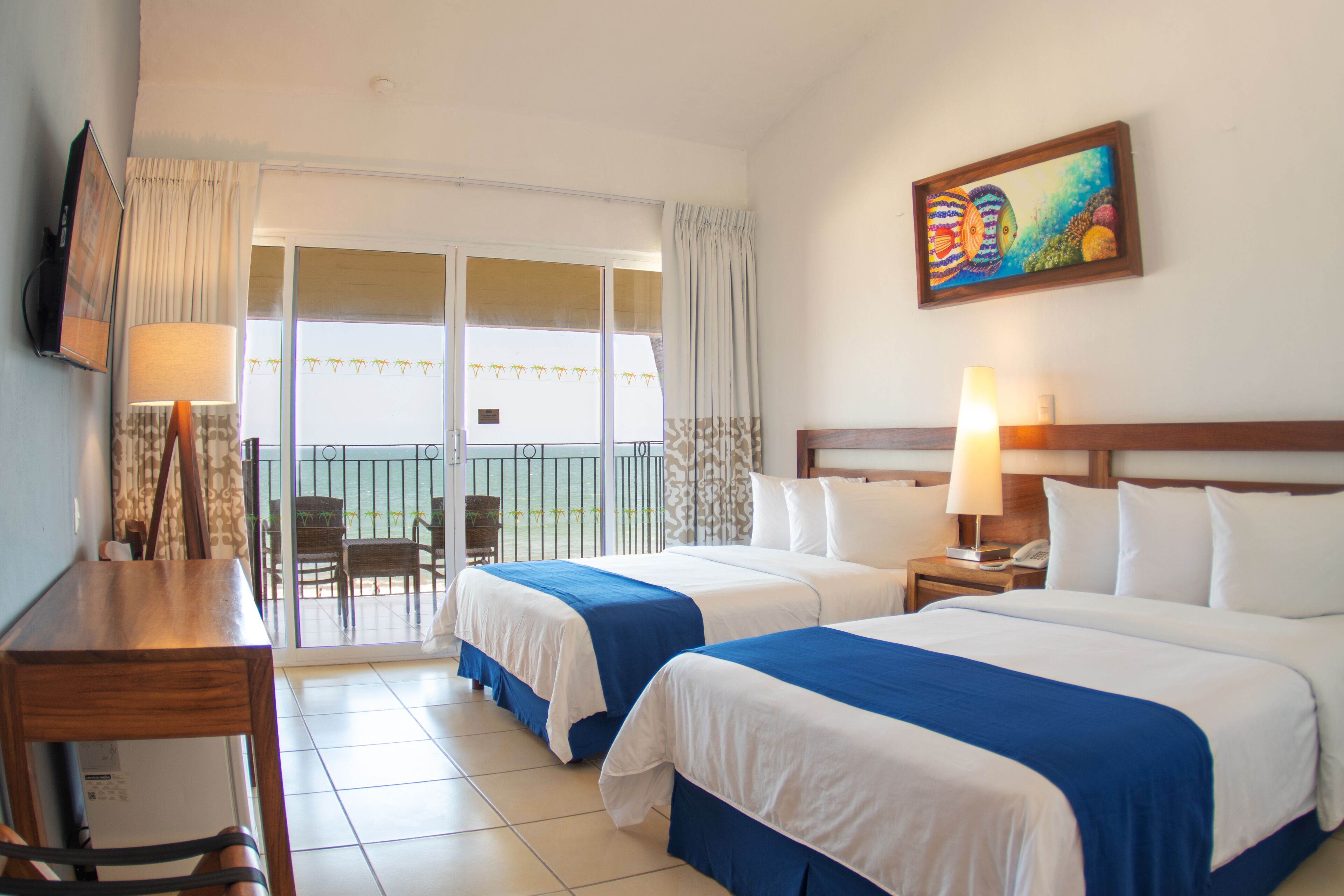 Habitación de hotel con dos camas, un escritorio, una silla, un balcón con vistas y un cuadro en la pared.
