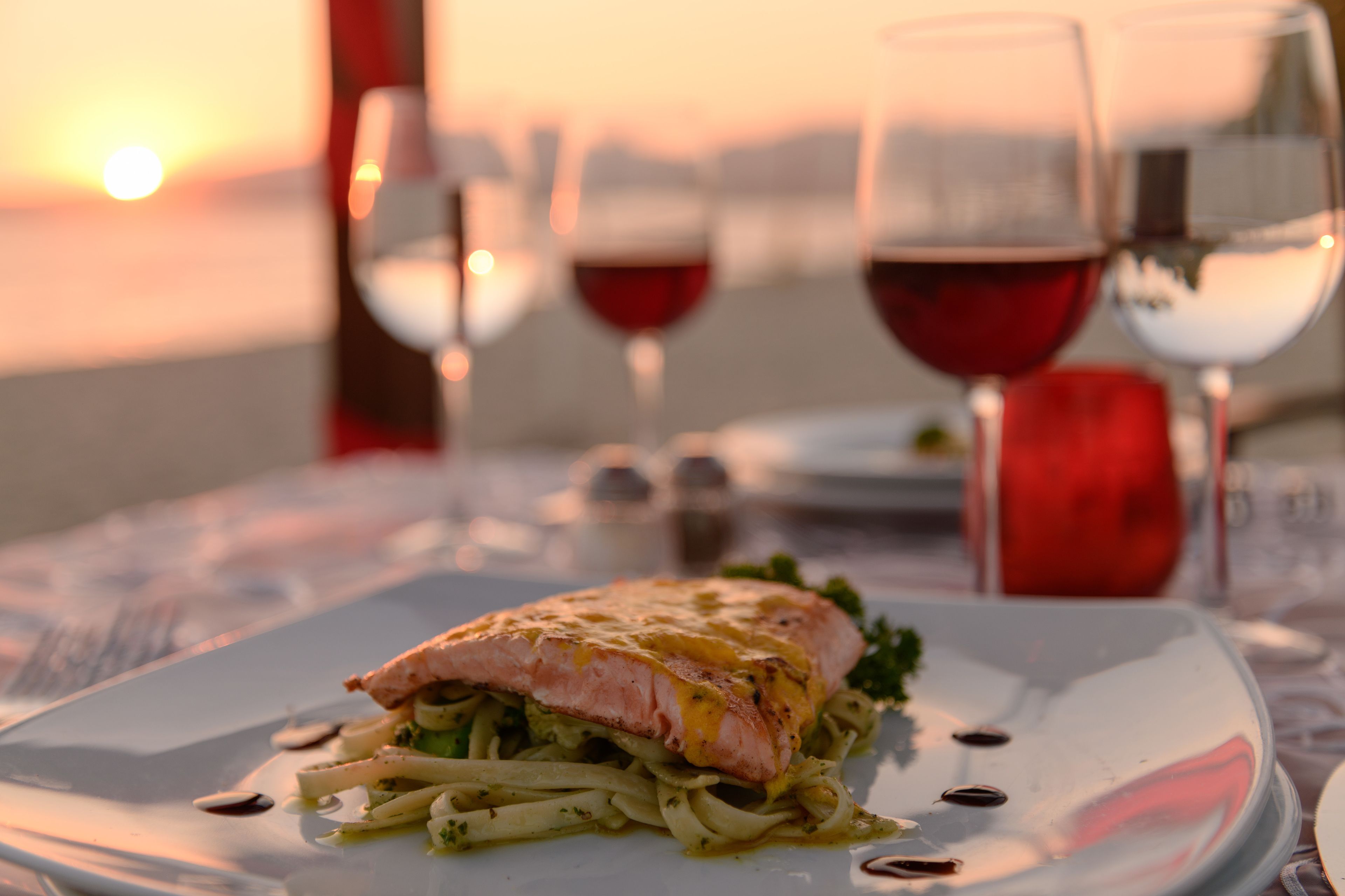 A meal with a piece of fish on a plate, accompanied by a glass of red wine, set against a sunset view.