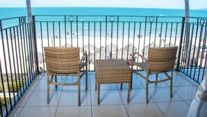 Un balcón con sillas de mimbre y una mesa, con vista a una playa con gente y palmeras.