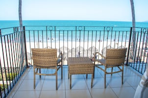 Un balcón con sillas de mimbre y una mesa, con vista a una playa con gente y palmeras.