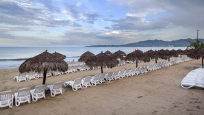Una playa con hileras de sillas de descanso blancas y sombrillas de paja, una orilla arenosa y vistas al mar y montañas distantes.