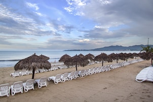 Una playa con hileras de sillas de descanso blancas y sombrillas de paja, una orilla arenosa y vistas al mar y montañas distantes.