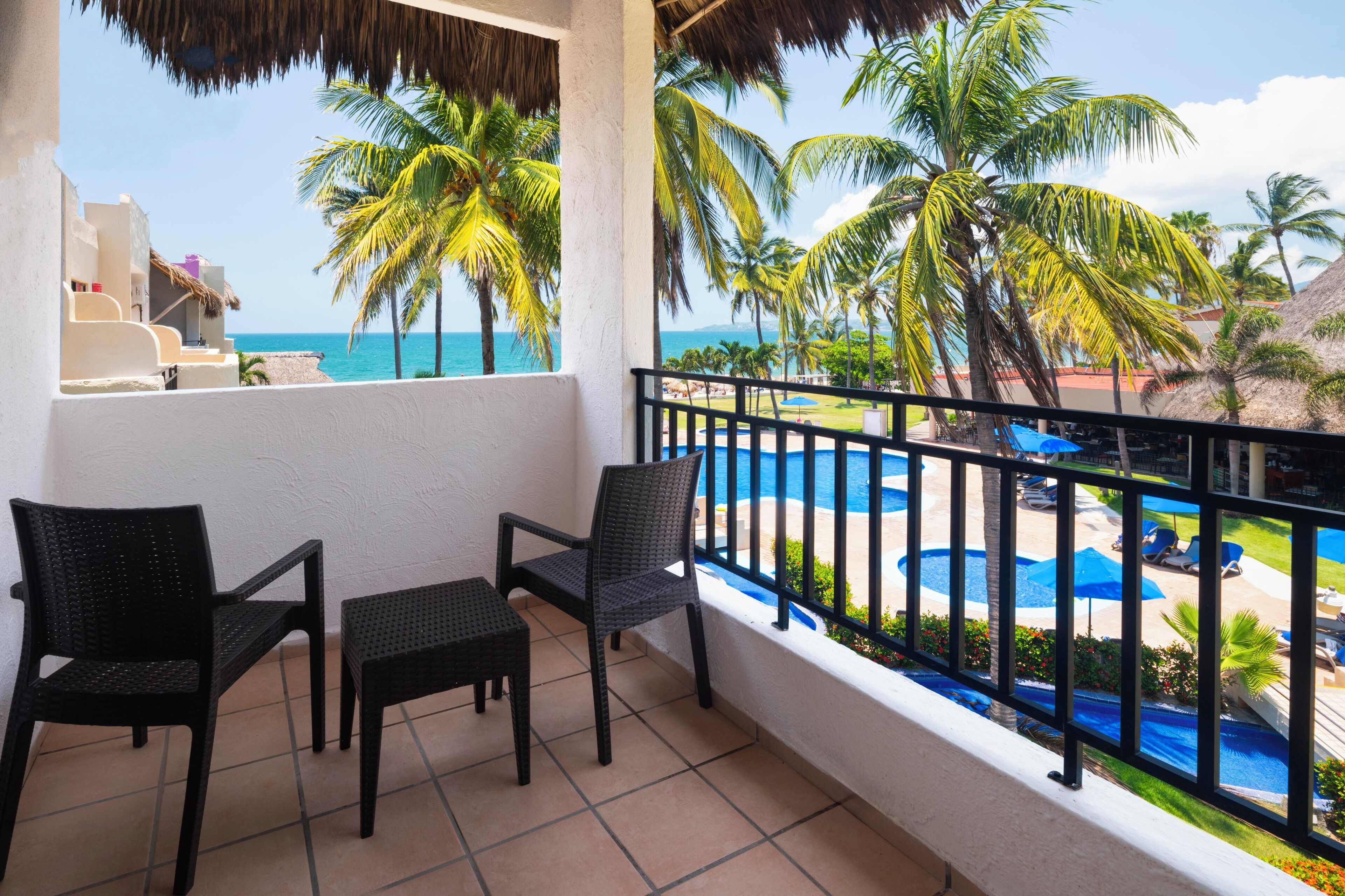 A balcony with two chairs and a small table, overlooking a pool area with palm trees and a view of the ocean.
