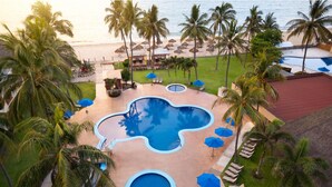 Zona de piscina en un resort, con varias piscinas, palmeras y sombrillas de playa.