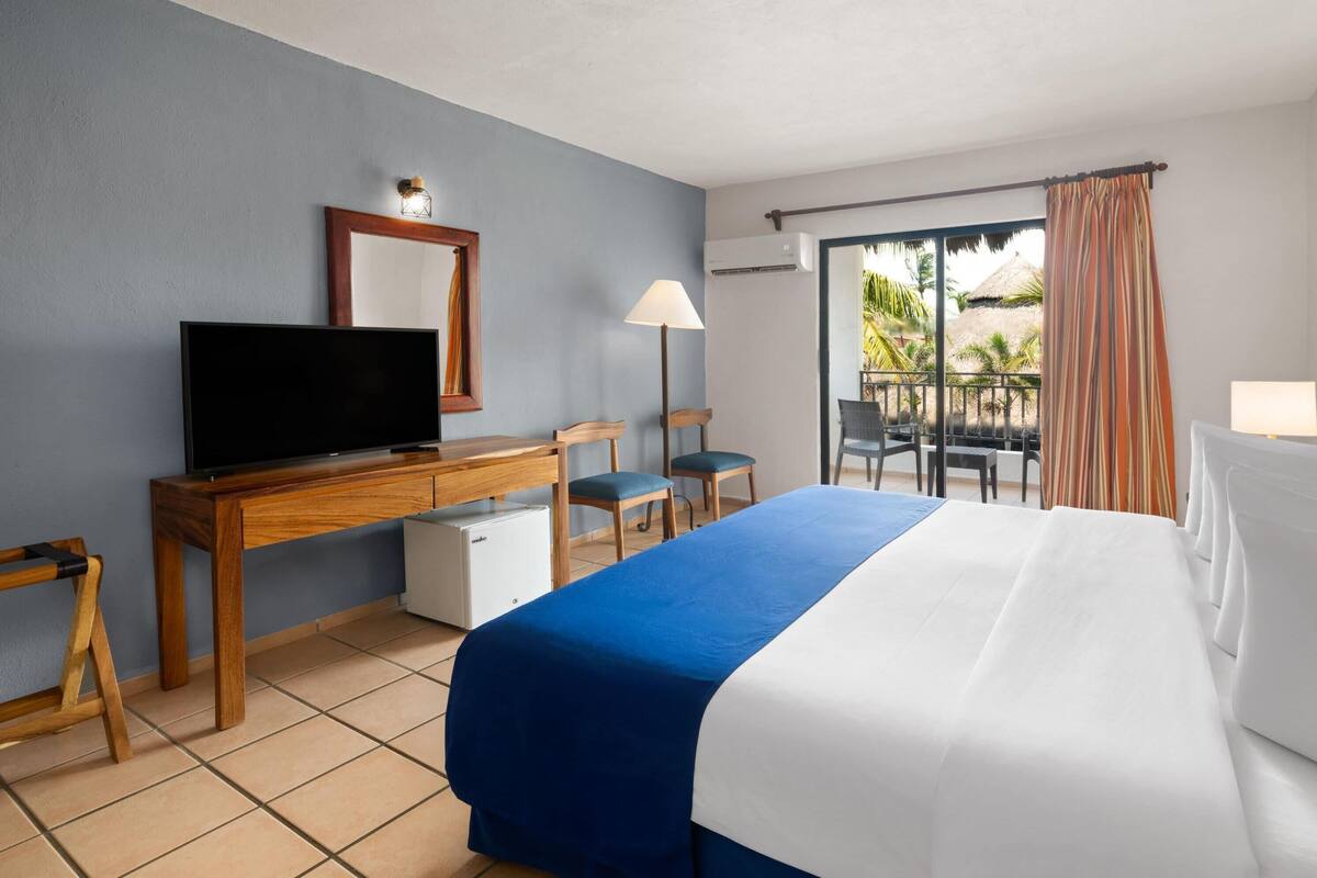 A hotel room with a bed, a desk with a television, a chair, and a balcony with a view of palm trees.
