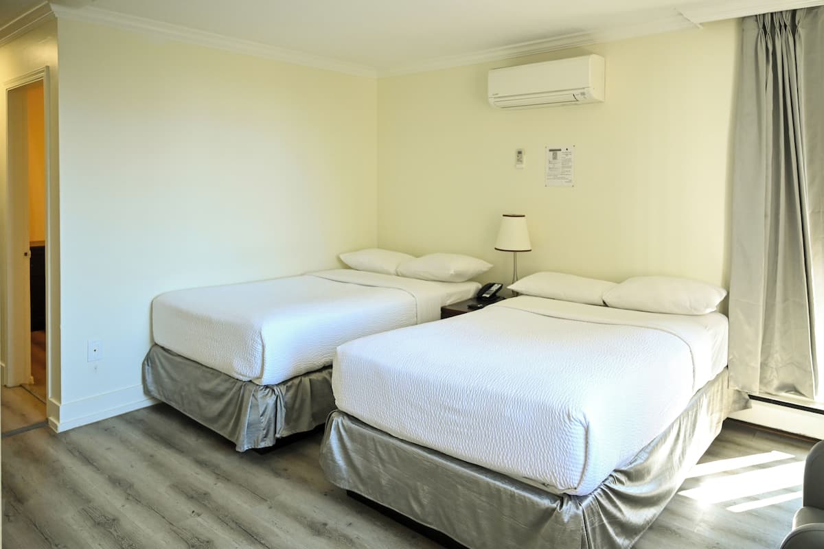 A hotel room with two beds, a bedside table with a lamp, an air conditioning unit, and a window with curtains.