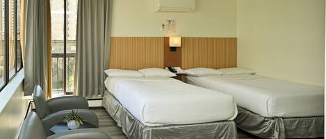 Habitación de hotel con dos camas, un escritorio, una silla, una planta en maceta y un ventanal con cortinas.