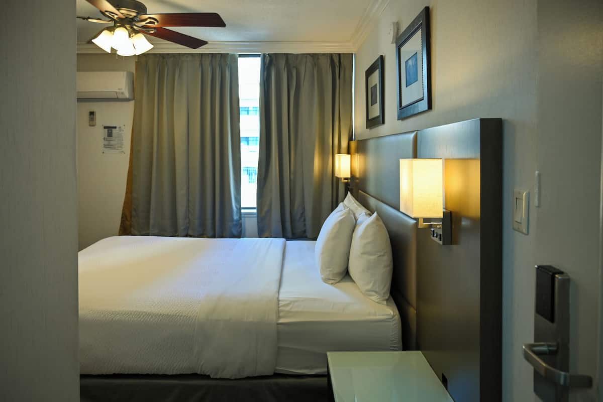 A hotel room with a bed, curtains, a ceiling fan, wall-mounted lights, and a door.