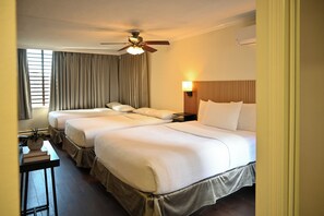 A hotel room with two beds, a ceiling fan, a bedside lamp, a window with curtains, and a small table with a plant and book.