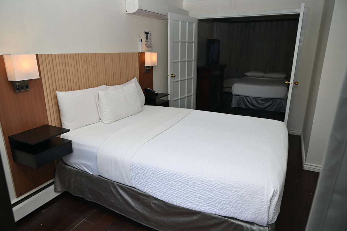 A hotel room with a large bed, bedside tables, a nightstand, a wall-mounted lamp, and a door leading to another room.