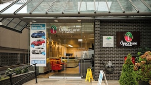 Exterior del Hotel Opportune con entrada de vidrio, un cartel para alquiler de coches independiente y un pequeño árbol de Navidad.