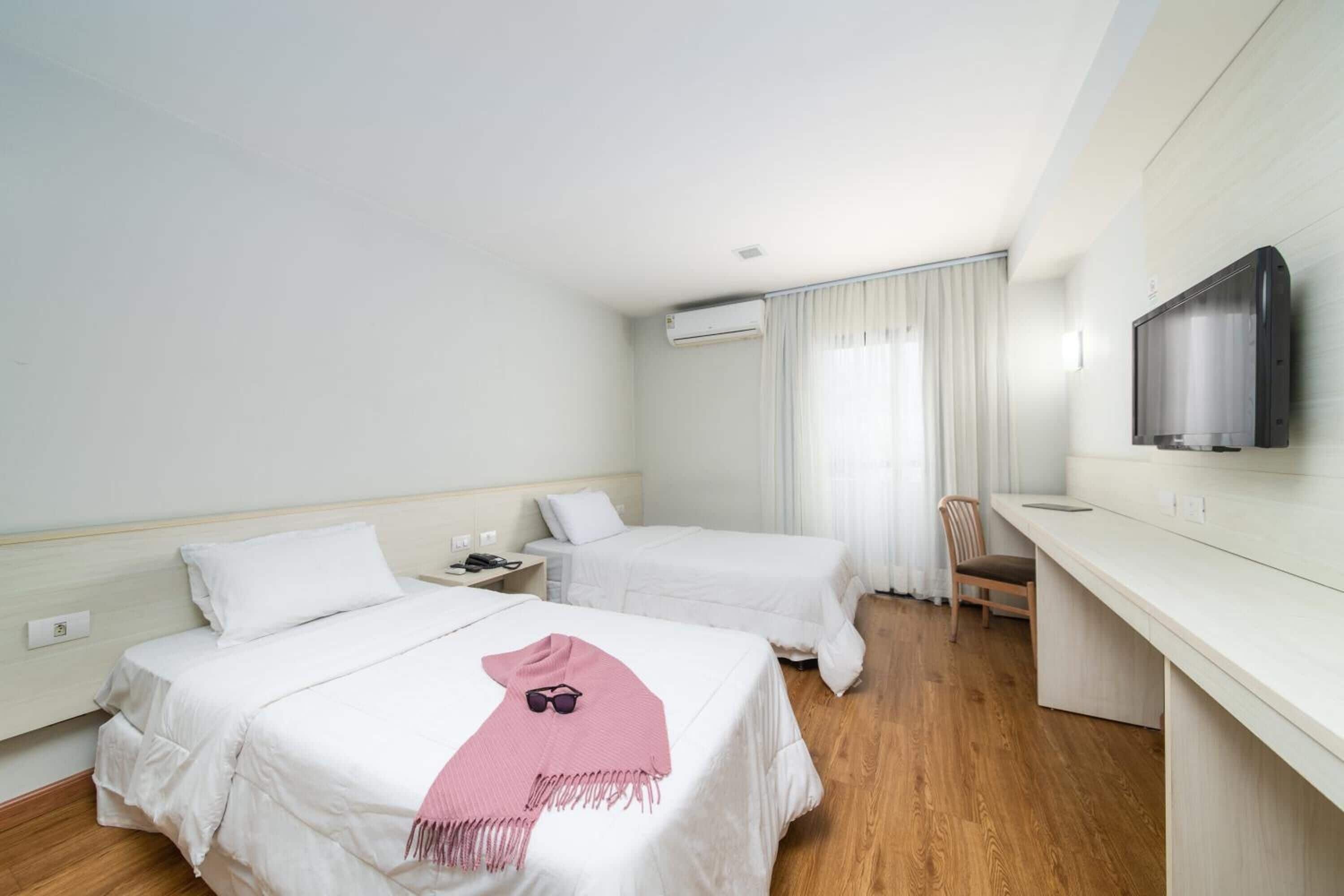 A hotel room with two beds, a desk, a chair, a TV, and an air conditioning unit.