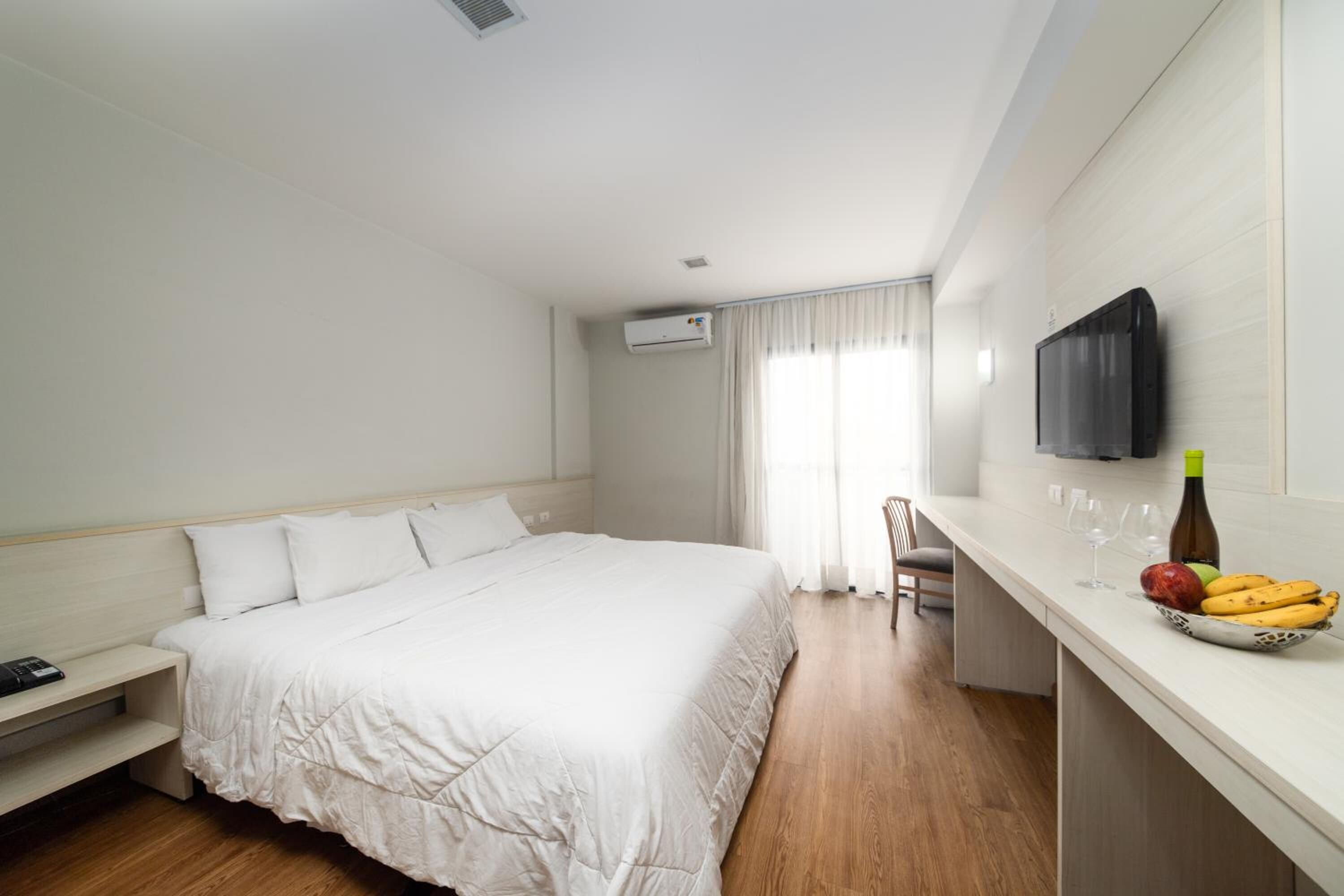 A hotel room with a bed, a TV mounted on the wall, a small table with fruit and a bottle, and a window with curtains.