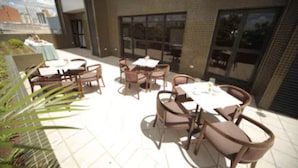 Un espacio de patio al aire libre con varias mesas y sillas, un pequeño jardín y un edificio con ventanas amplias y fachada de ladrillo.