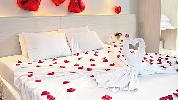Una habitación de hotel con una cama bien hecha, ropa de cama blanca, pétalos de rosa y decoraciones con forma de corazón en la pared.