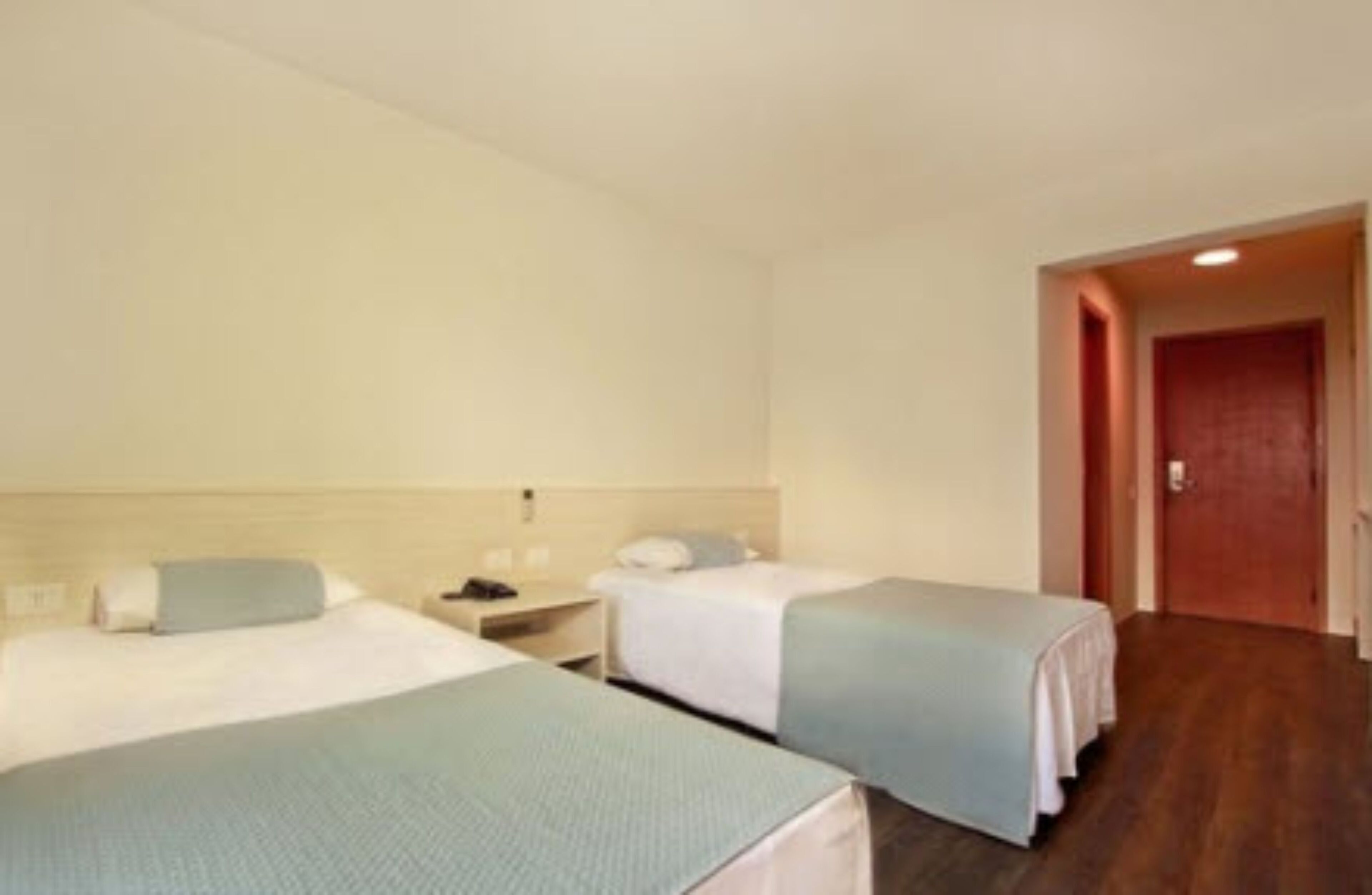 A hotel room with two single beds, a nightstand, a wooden floor, and a door leading to another room.