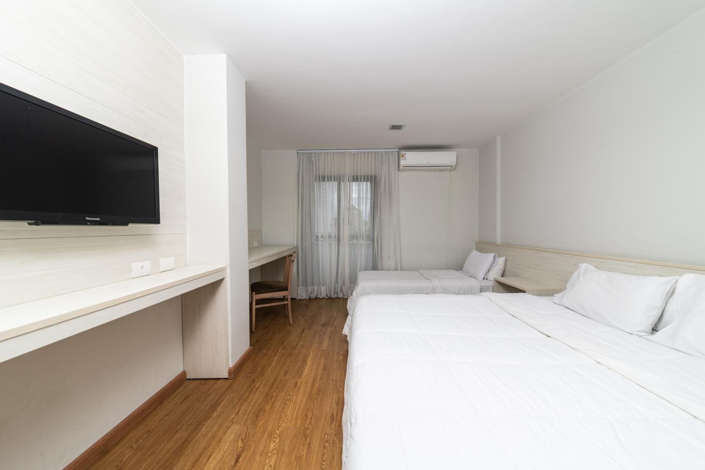 A hotel room with a flat-screen TV, a desk, two beds, a window with curtains, and an air conditioning unit.