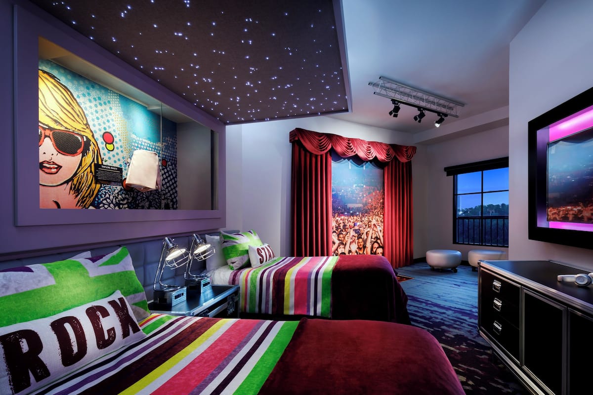 A hotel room with a starry ceiling, a large bed with a colorful striped blanket, a flat-screen TV, and a cityscape view through the window.