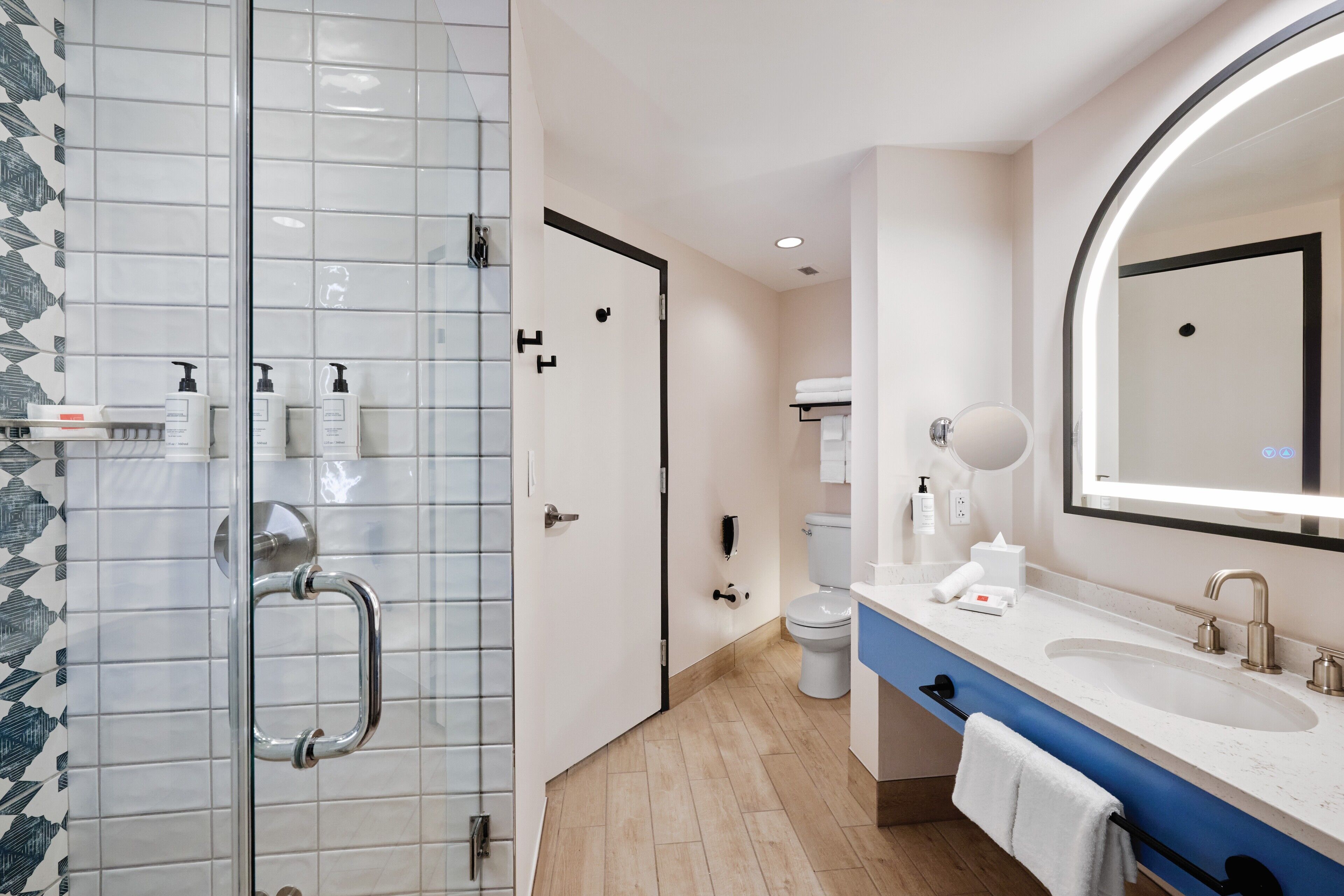 Een moderne badkamer met een glazen douchecabine, een wit toilet, een blauwe wastafel met twee wasbakken en een grote spiegel.