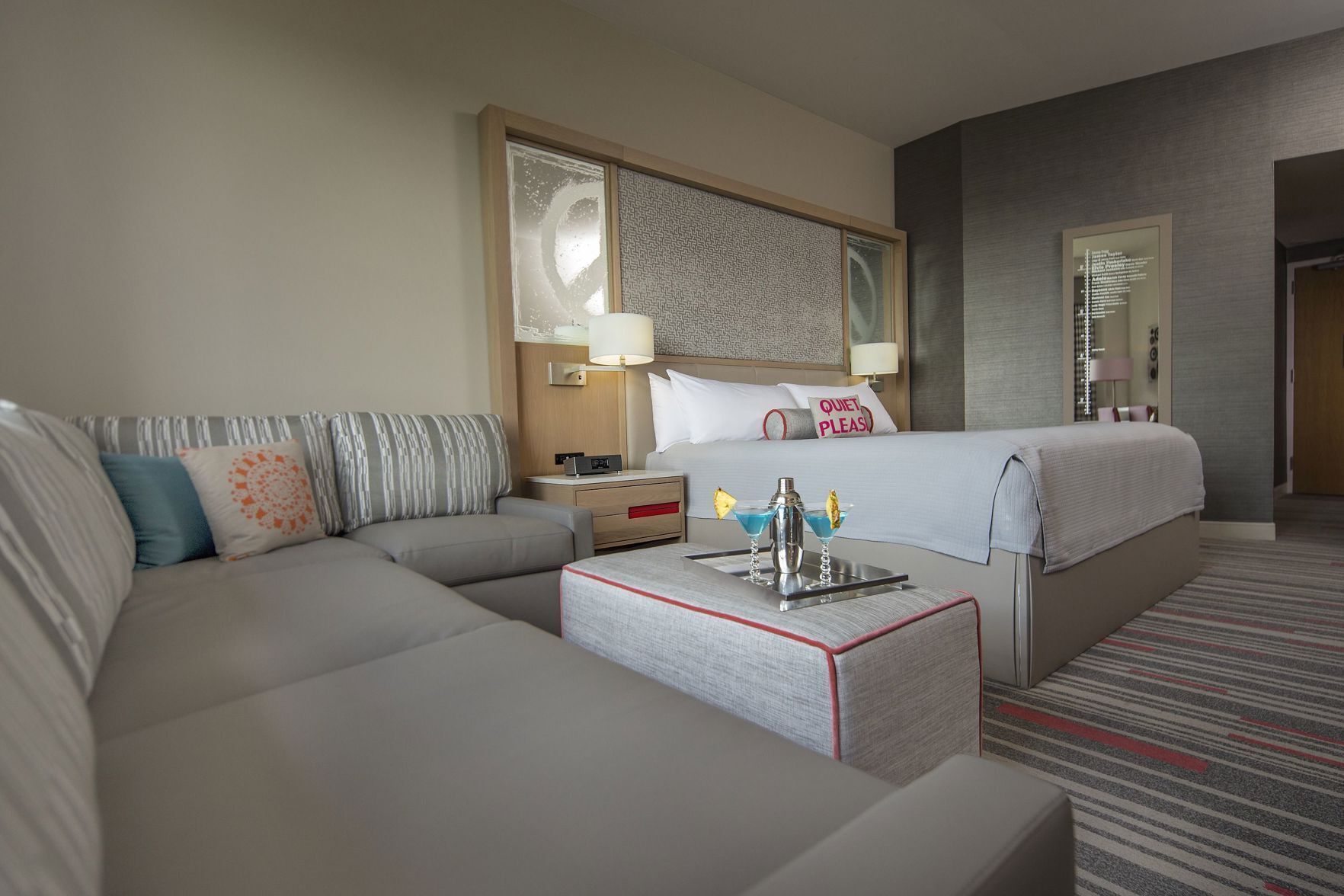 A hotel room with a bed, a sofa, a bedside table with a lamp, a tray with two drinks, and a wall with a mirror and a clock.