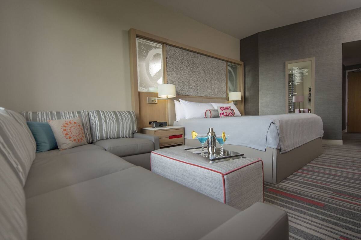 A hotel room with a bed, a sofa, a bedside table with a lamp, a tray with two drinks, and a wall with a mirror and a clock.